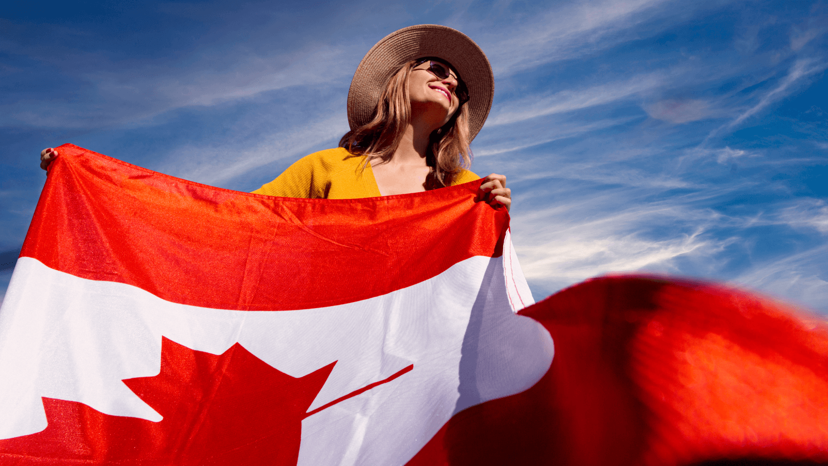 Cheerful woman proudly waving a Canadian flag in front of a vibrant blue sky.