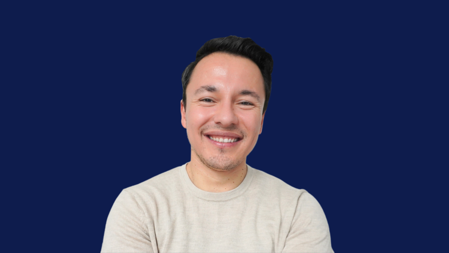 A smiling man with short dark hair wearing a light beige crewneck t-shirt, set against a solid dark blue background.